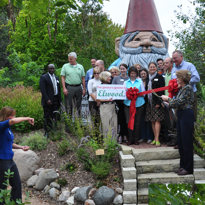 Gnome Gname Reveal - a group stands in front of Elwood with a sign which reads, "The gnome's name is Elwood." A woman stands in front of a red ribbon with large scissors, about to cut.