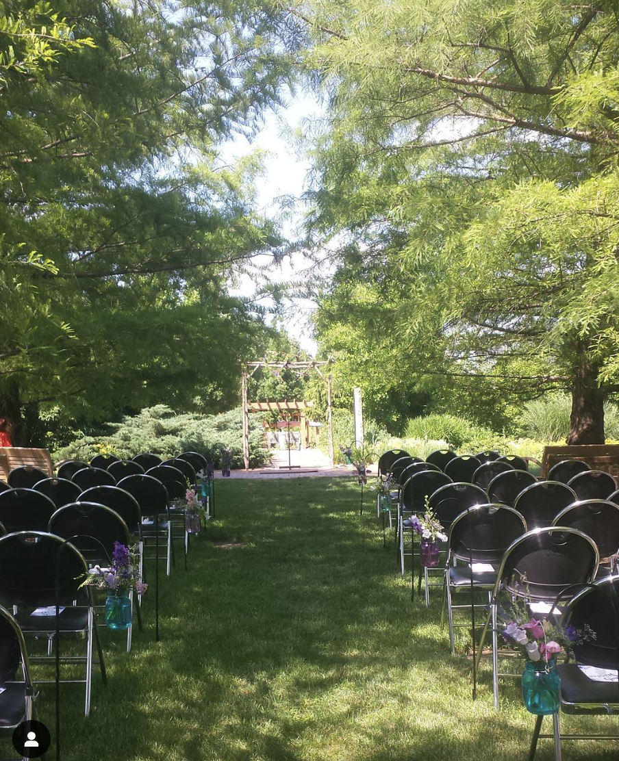 Looking down the aisle of an outdoor wedding space at Reiman Gardens. Trees line the exterior of the shot, flanked by rows of chairs. Down the aisle, an arch awaits the bride and groom.
