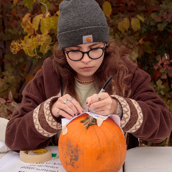 A woman wearing warm autumn clothes traces a design onto a pumpkin using a paper stencil.