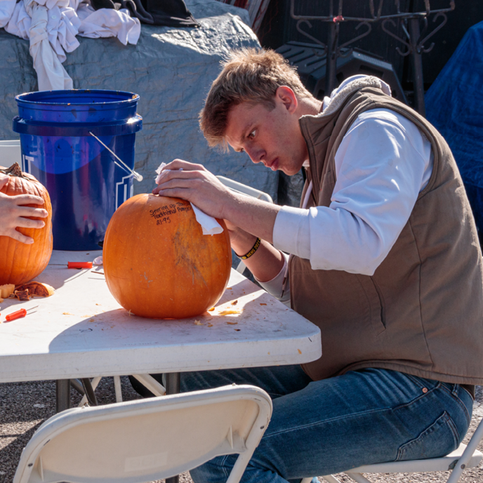 A college-aged man holds a pumpkin steady as he hunches over it to carve.