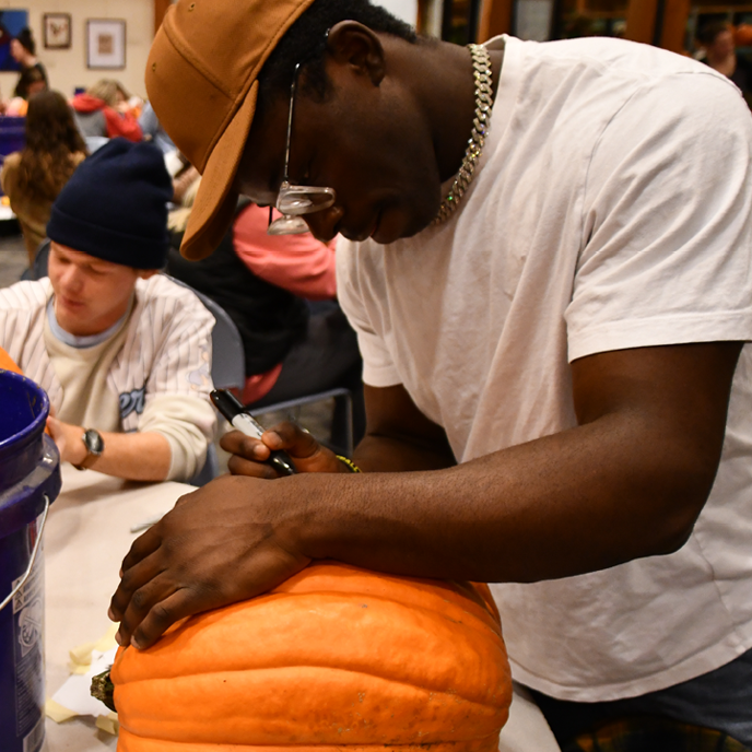 A college-aged man holds a pumpkin steady as he hunches over it to stencil.
