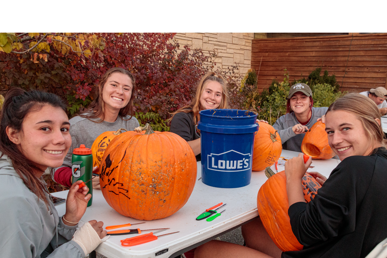 A group of college-aged women carve pumpkins together.
