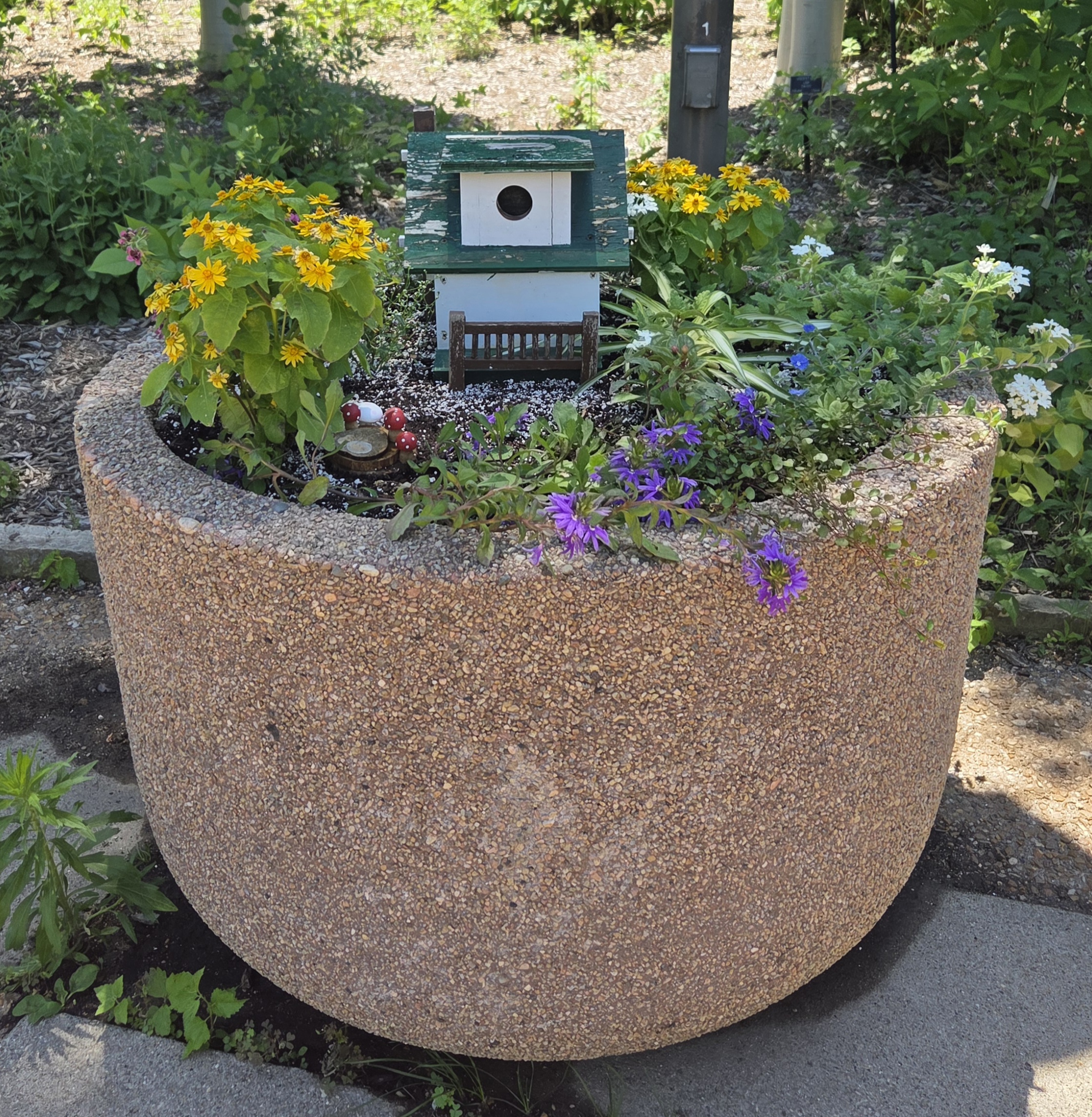 A container with a white and green birdhouse along with multiple plants