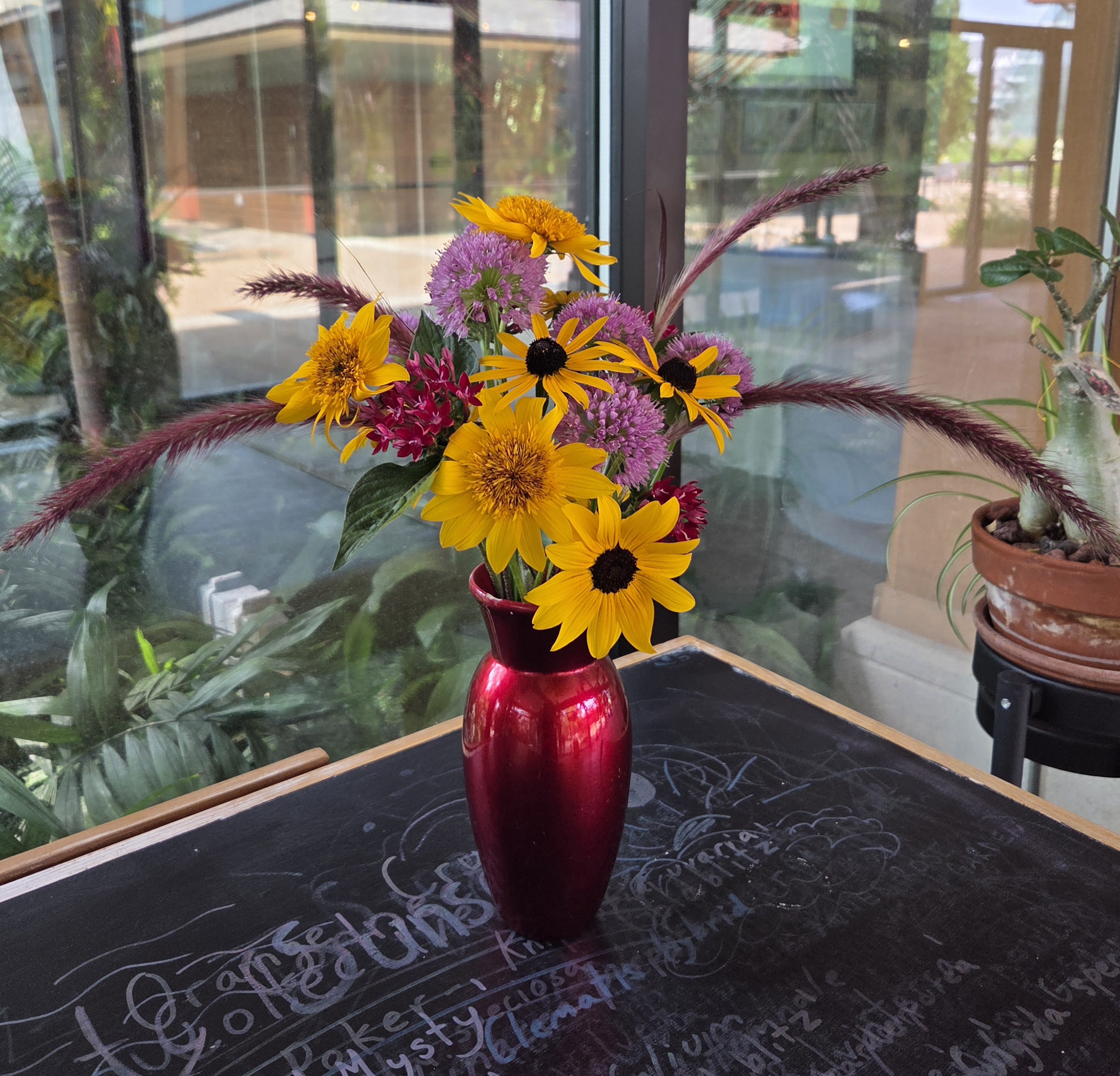 A red vase with an assortment of yellow and purple flowers along with stems of purple grass