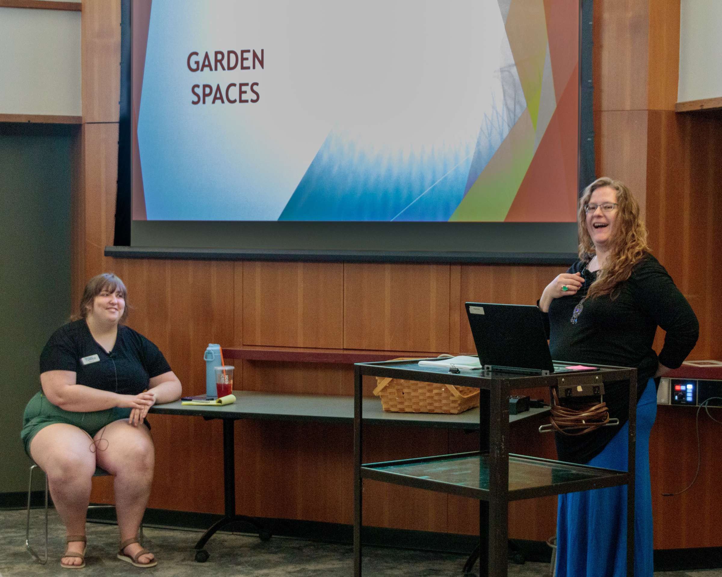 Interior of auditorium, two women stand before a screen, about to give a presentation on Garden Spaces.