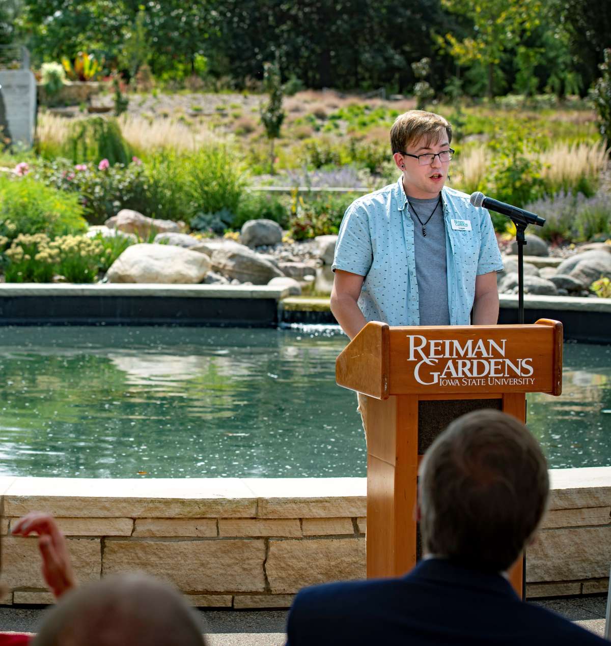 Before the reflection pool at Sycamore Falls, Logan Metzger, a student worker from Reiman Gardens, speaks at a podium before a crowd. 