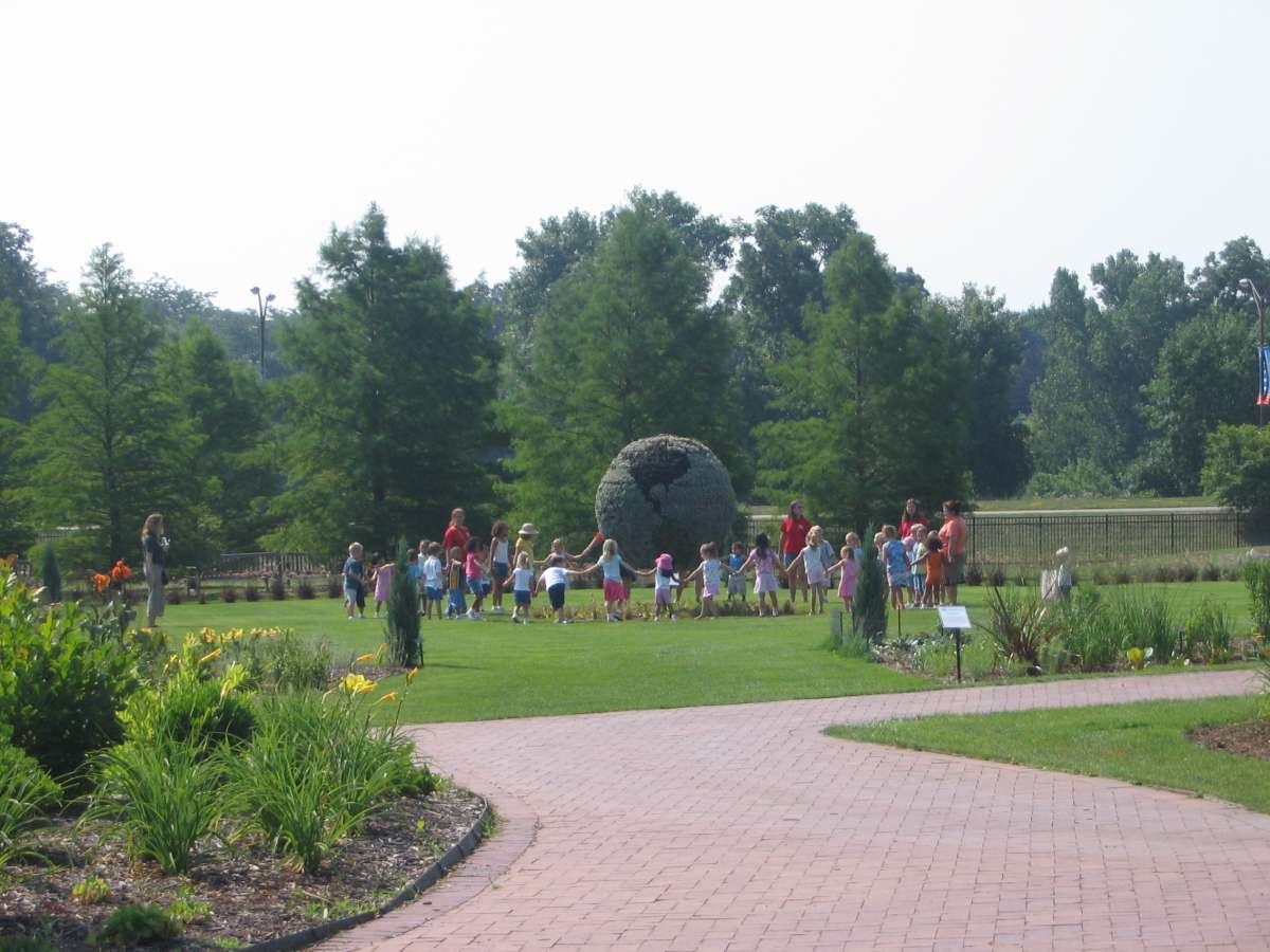a group of schoolchildren and adults linking hands around a giant topiary globe in a garden