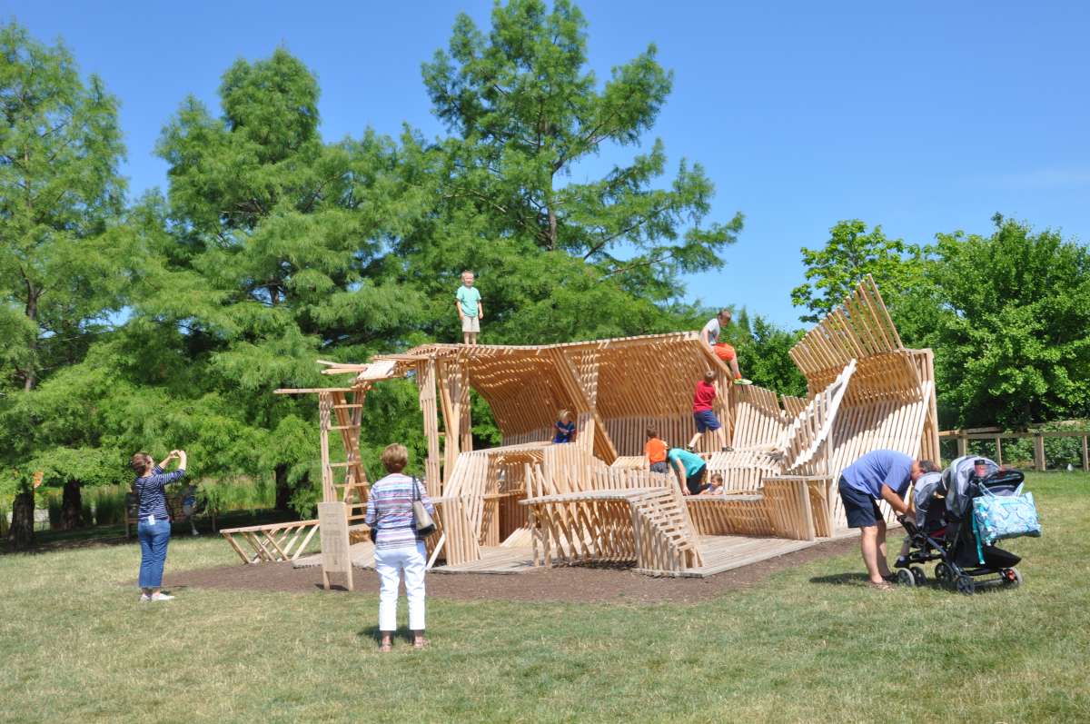 a large wooden play structure made from 2x2 pieces of wood set in a garden, with people of various ages playing on and around it