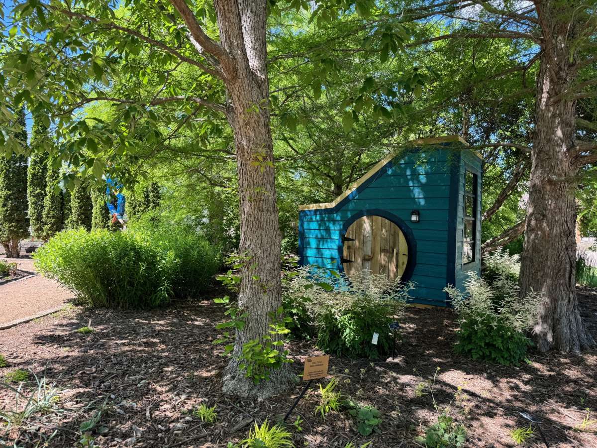 a dark teal tiny house with a moss-covered sloped roof set into a garden space under a redwood tree