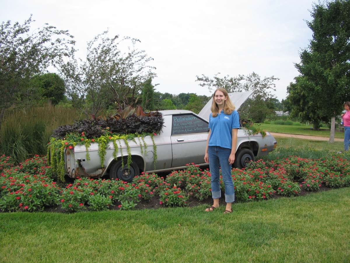 a silver truck in a garden, planted with plants, behind a young white female