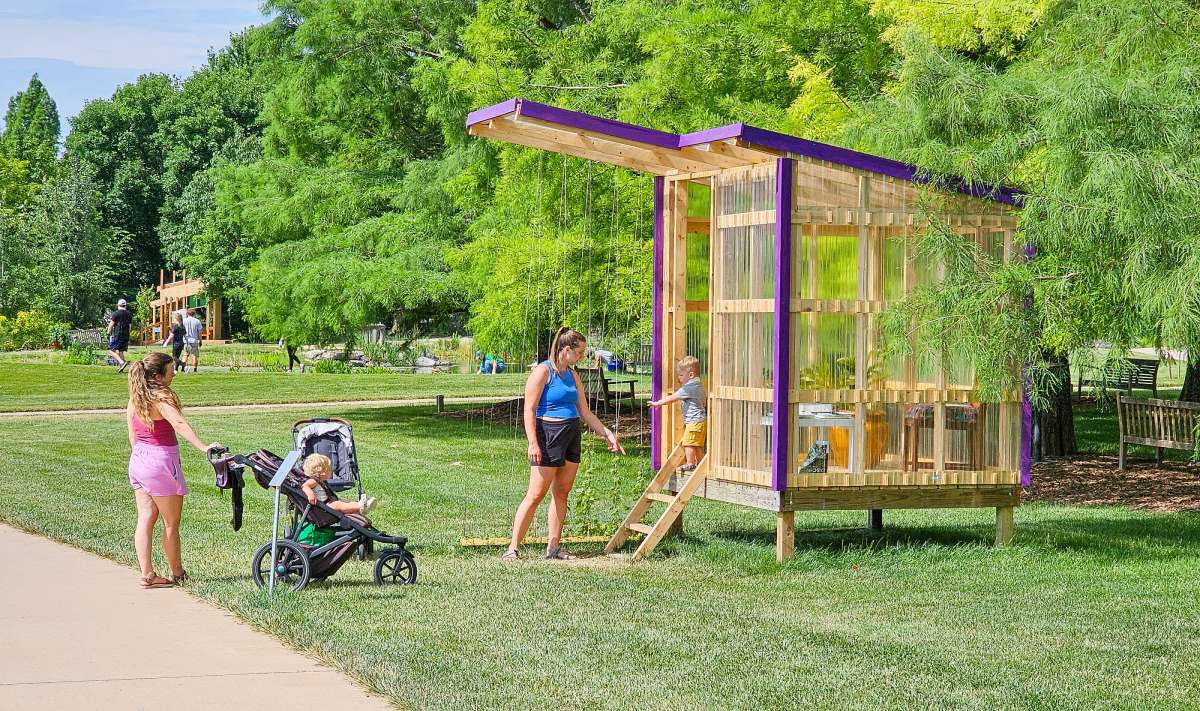 a greenhouse-inspired playhouse in front of a row of trees in a garden, with people in and around it