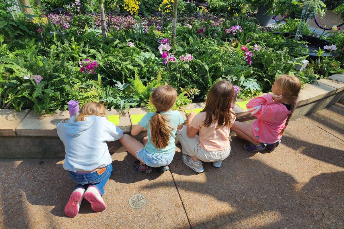 4 Girls sitting on the ground in the conservatory facing away from the camera writing on half sheets of paper.