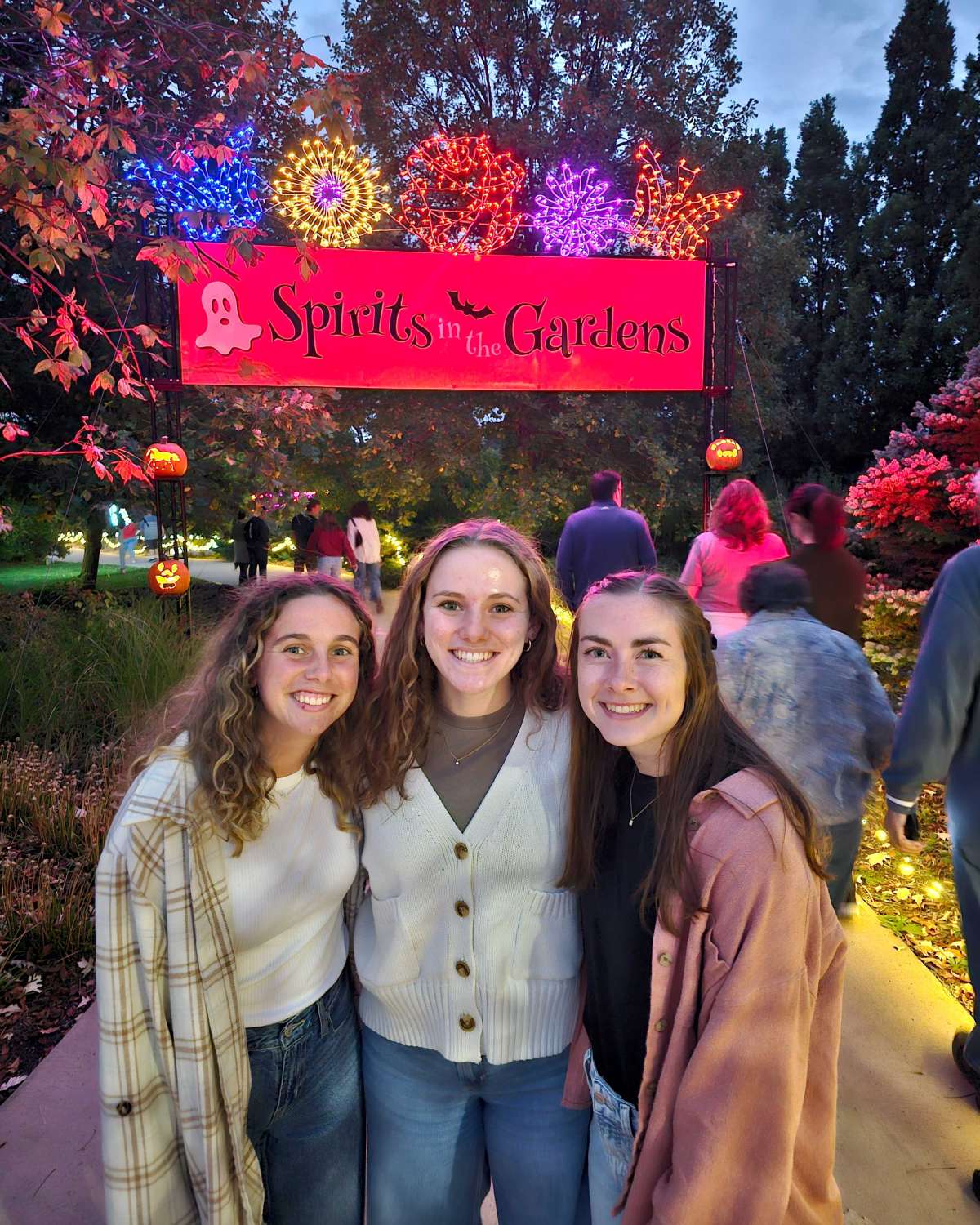 Three young women smiling in front of a "Spirits in the Gardens" Halloween event sign, surrounded by people and festive lights in an outdoor garden setting at dusk.