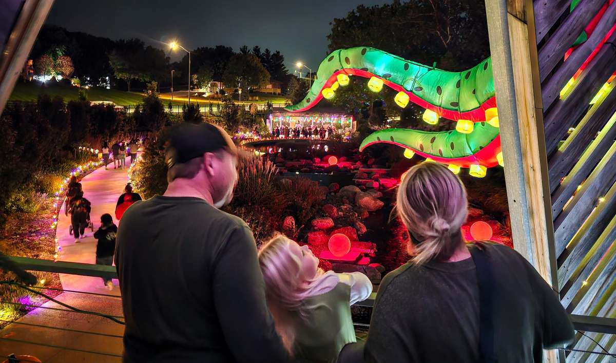 Three people stand on a wooden platform overlooking a brightly lit nighttime outdoor scene with colorful lanterns shaped like a large serpent and a crowd walking along a path.