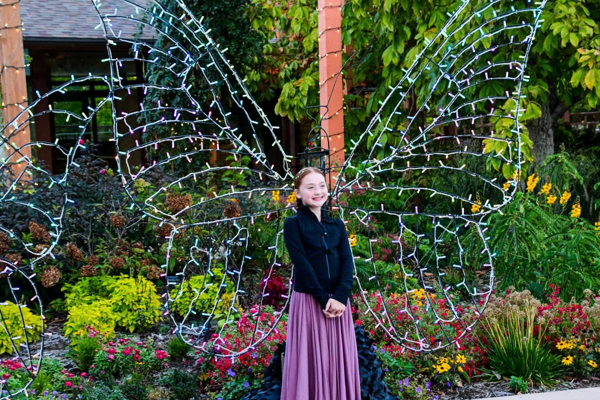A young girl wearing a long skirt and dark jacket stands smiling in front of a large butterfly-shaped light decoration surrounded by colorful flowers and greenery.