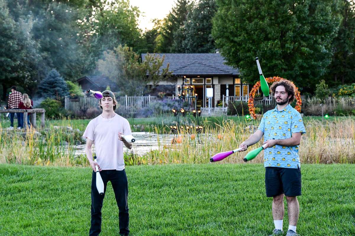Two adults stand on grass near a pond in a park-like setting while juggling clubs, with one person wearing a light pink shirt and the other wearing a blue patterned shirt.