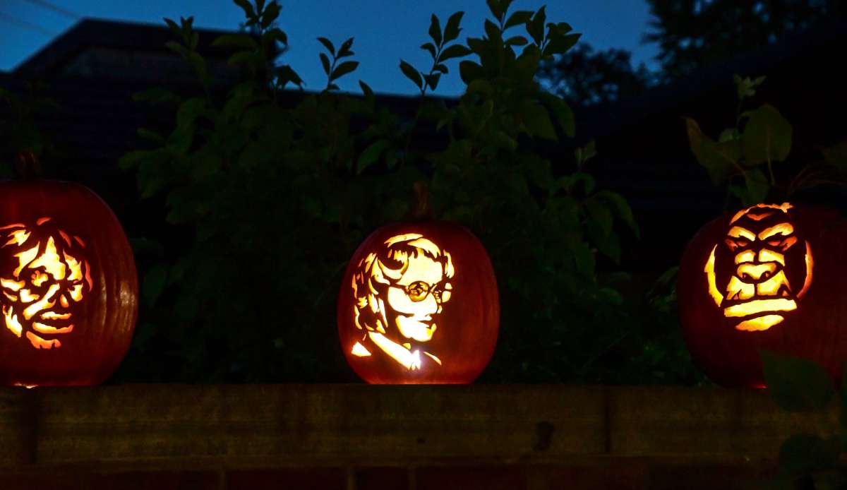 Three intricately carved pumpkins with illuminated faces are displayed outdoors against a dark evening sky.