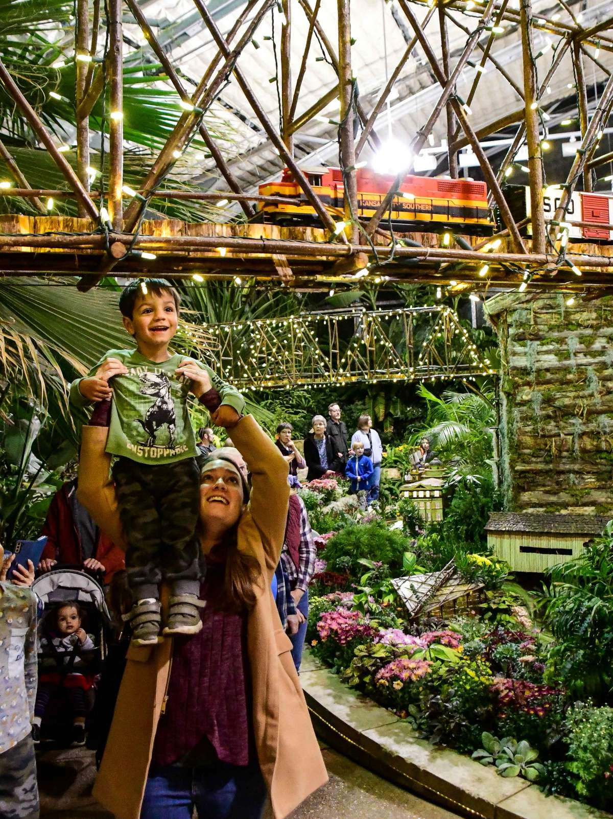 Woman holding a young boy up to see a miniature train on a bridge in a conservatory garden.