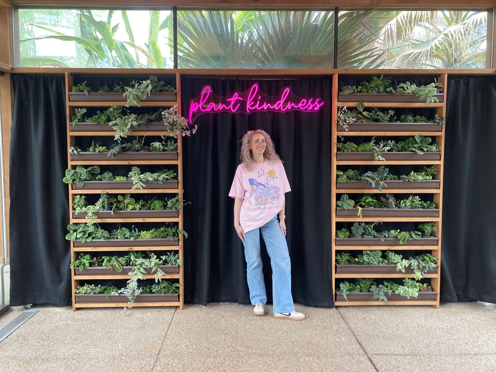 plants in boxes along the wall on either side of a person standing under a neon sign that says Plant Kindness